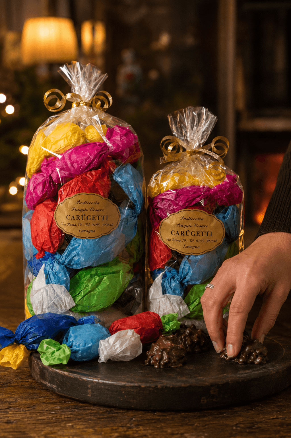 Due bundle Carugetti La Tavola in atmosfera natalizia, una mano sceglie una meringa al cioccolato fondente accanto alle confezioni con fiocco dorato